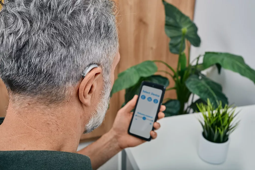 A mature man with hearing loss adjusts settings for his BTE Bluetooth hearing aid via his smartphone app. 