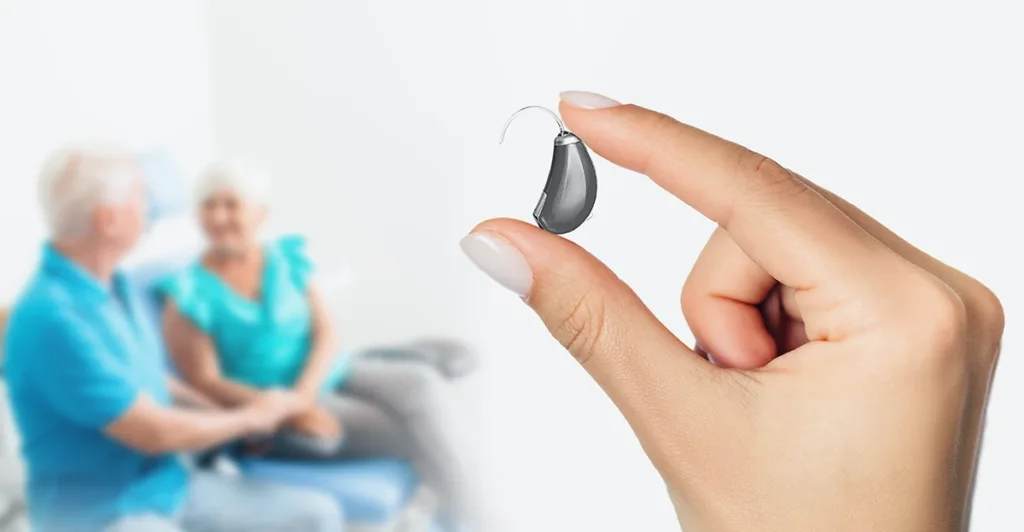 Hearing health specialist holding up ITE hearing aid, with two older adult people out of focus in the background, representing how hearing aids reduce the risk of dementia in older adults, seniors. 